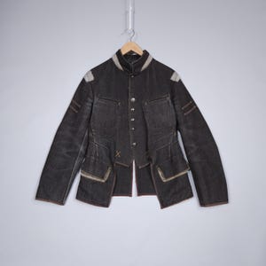 May include: A dark grey denim jacket with a distinctive design. The jacket has a stand-up collar, multiple pockets, and decorative stitching. It features a button-up front and brown trim. The jacket is suspended from a wooden hanger.