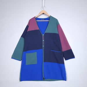 Pode incluir: Um cardigan longo com blocos de cores em tons de azul, verde e roxo. O suéter tem decote em V, mangas compridas e um único bolso. Está abotoado e pendurado em um cabide de madeira.