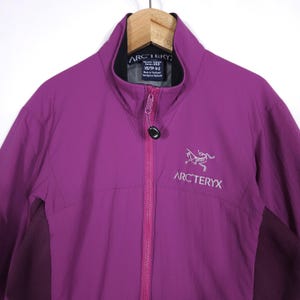 May include: A vibrant purple jacket with a full-length zipper and a high collar. The jacket features the Arc'teryx logo in white on the chest. The jacket is hanging on a wooden hanger.