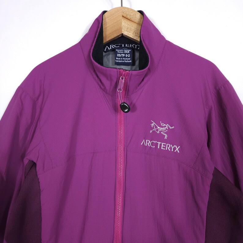 May include: A vibrant purple jacket with a full-length zipper and a high collar. The jacket features the Arc'teryx logo in white on the chest. The jacket is hanging on a wooden hanger.