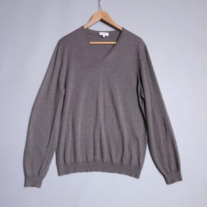 May include: A long-sleeved, V-neck sweater in a muted brown color. The sweater is hanging on a wooden hanger against a gray background. The neckline and cuffs are ribbed.