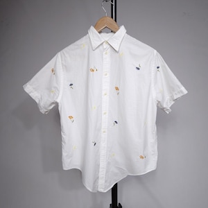 Puede incluir: Camisa blanca de manga corta con bordados florales amarillos, azules y naranjas. La camisa está abotonada y colgada de una percha de madera. Una prenda casual para el día a día.