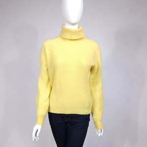 May include: A pale yellow turtleneck sweater with long sleeves. The sweater has a ribbed texture and a relaxed fit. The garment is displayed on a mannequin. The sweater is a soft, warm, and comfortable garment.