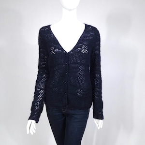 May include: A navy blue, open-weave cardigan with a V-neck and button closure. The long-sleeved sweater is displayed on a mannequin, paired with dark blue jeans. The knitwear has a delicate, airy design.
