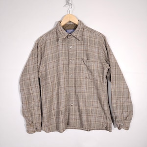 Puede incluir: Una camisa de franela a cuadros marrón y beige con cierre de botones y un solo bolsillo en el pecho. La camisa tiene mangas largas y cuello.