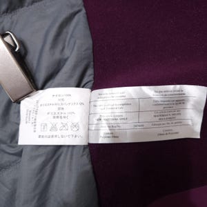 May include: Close-up of garment tags with Japanese and English text. The tags are white with black text and are attached to a purple and gray fabric. One tag states "NEW MATERIAL ONLY".