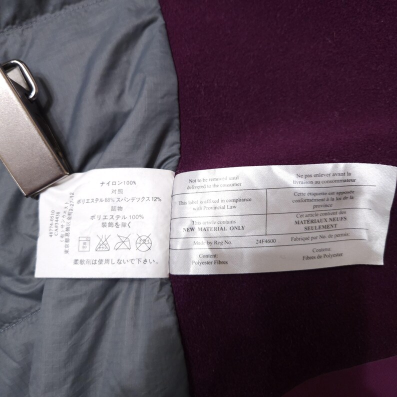May include: Close-up of garment tags with Japanese and English text. The tags are white with black text and are attached to a purple and gray fabric. One tag states "NEW MATERIAL ONLY".