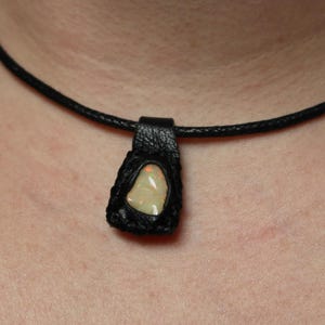 May include: A black leather pendant necklace featuring a light-colored, irregularly shaped stone. The stone is set within a black leather frame and hangs from a thin, black cord. The necklace is displayed against a skin tone background.