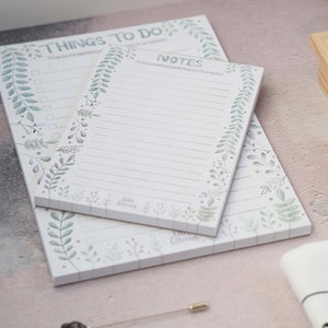 Puede incluir: Dos juegos de bloc de notas blancos con bordes florales verdes. El bloc de notas superior tiene el texto "Things to Do" y el bloc de notas inferior tiene el texto "Notes".