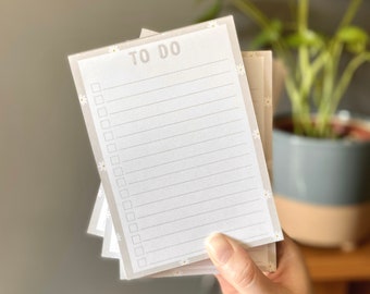 May include: A stack of to-do list notepads with a white background and a light gray border. The notepads are decorated with small white daisies. The top notepad has the words "TO DO" printed in black.