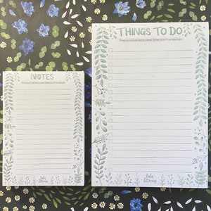 May include: Two white notepads with green floral borders. The top notepad has the text "THINGS TO DO: Some days I am the flower, some days I am the rain." The bottom notepad has the text "NOTES: Sometimes I am the flower, sometimes I am the rain."