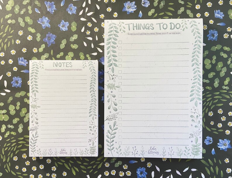 May include: Two white notepads with green floral borders. The top notepad has the text "THINGS TO DO: Some days I am the flower, some days I am the rain." The bottom notepad has the text "NOTES: Sometimes I am the flower, sometimes I am the rain."
