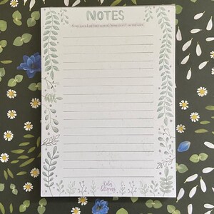 May include: A white notepad with a green floral border and the text "NOTES" at the top. The quote "Some days I am the flower, some days I am the rain." is printed below the title. The bottom of the notepad has the text "Kalm lettering."