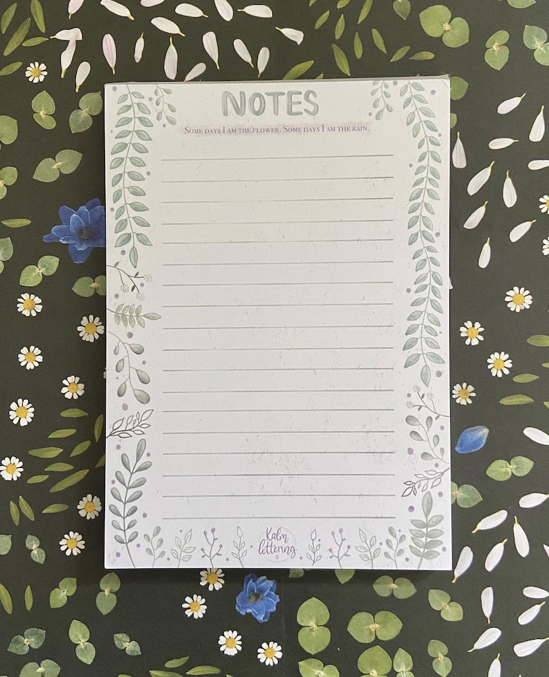 May include: A white notepad with a green floral border and the text "NOTES" at the top. The quote "Some days I am the flower, some days I am the rain." is printed below the title. The bottom of the notepad has the text "Kalm lettering."