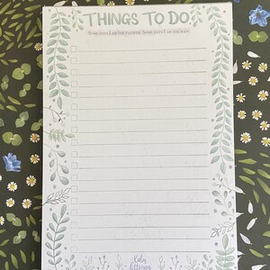 May include: A white notepad with a green floral border and the text "THINGS TO DO" at the top. The notepad has a quote that reads "SOME DAYS I AM THE FLOWER. SOME DAYS I AM THE RAIN." and a list of blank lines with checkboxes. The bottom of the notepad has the text "Kalm lettering."