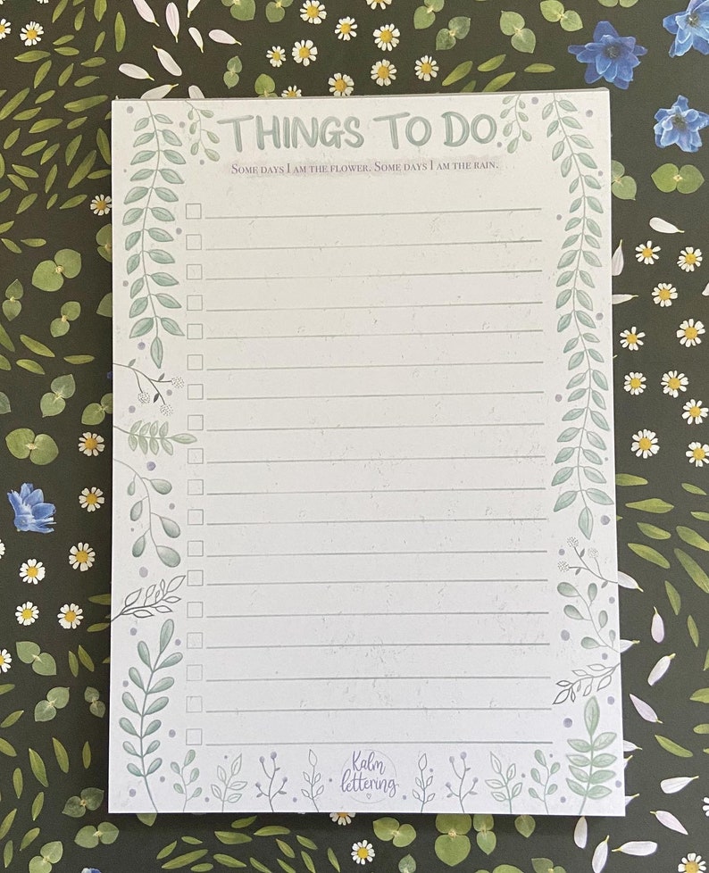 May include: A white notepad with a green floral border and the text "THINGS TO DO" at the top. The notepad has a quote that reads "SOME DAYS I AM THE FLOWER. SOME DAYS I AM THE RAIN." and a list of blank lines with checkboxes. The bottom of the notepad has the text "Kalm lettering."
