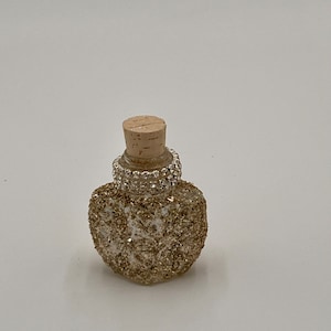 May include: A small, clear glass bottle with a cork stopper and a gold glitter finish. The bottle is decorated with a band of rhinestones around the neck.