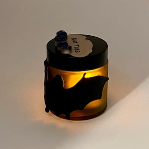 May include: A glass jar with a black lid and a bat silhouette on the front. The jar is filled with a yellow liquid and has a label that says "Bat Toos".