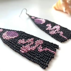 Peut inclure: Une paire de boucles d'oreilles perlées faites à la main. Les boucles d'oreilles sont noires avec un motif rose et violet. Le motif comprend un croissant de lune et un motif floral. Les boucles d'oreilles ont des crochets argentés.