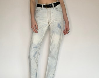 May include: Light blue denim jeans with a faded, tie-dye effect. The jeans have a high waist and a straight leg fit. The model is wearing a black crop top and black sandals.