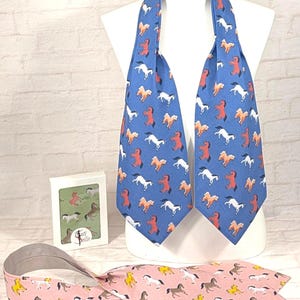 May include: A blue ascot and a pink tie, both patterned with horses, are displayed on a white mannequin. The ascot features white, red, and tan horses. The tie has brown and yellow horses. A small box with horse illustrations is also present.