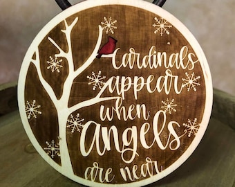 May include: A wooden Christmas ornament shaped like a ball with a brown background and white snowflakes. A white tree with a red cardinal is in the center. The text "Cardinals appear when angels are near" is written in white on the ornament.