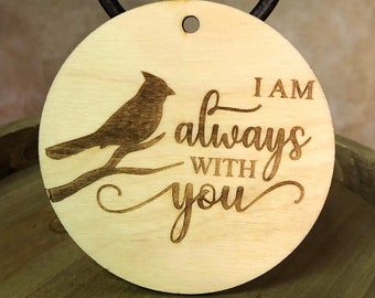 May include: A round, wooden pendant with a dark brown cardinal bird design perched on a branch. The text "I AM always WITH you" is etched into the light wood. A black leather cord is attached for wearing.