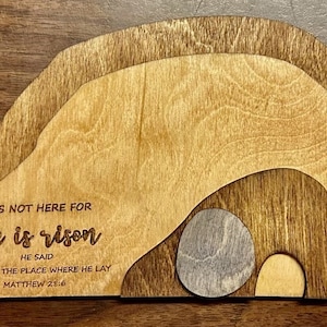 Può includere: Scena pasquale in legno con design a strati. Il testo "He is risen" è inciso sul legno di colore chiaro. La scena raffigura un sepolcro vuoto con una pietra a forma di uovo. È inclusa anche la scrittura Matteo 28:6.