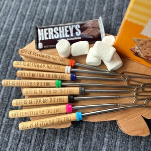 May include: A set of eight telescopic roasting sticks with wooden handles engraved with the text "LIFE IS BETTER BY THE FIRE" and colorful silicone rings, accompanied by a Hershey's milk chocolate bar and white marshmallows.