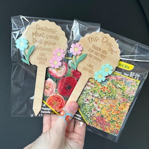 May include: Two wooden flower-shaped garden stakes with text, "Teachers plant seeds that grow forever" and "Thank you for helping me grow." Each stake has decorative flower accents. Packets of flower seeds are visible.