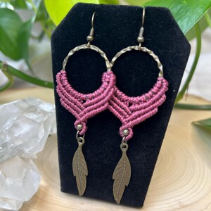 May include: A pair of dangle earrings featuring a hammered bronze-tone metal hoop at the top, accented with mauve-colored macrame cord knotted in a chevron pattern, and finished with a small bronze-tone metal feather charm hanging from the center.