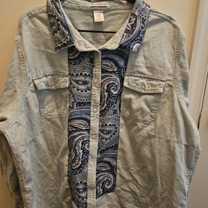 Könnte beinhalten: Ein hellblaues Jeanshemd mit Knopfleiste und einem dunkelblau-weißen Paisley-Muster auf der Vorderseite und am Kragen. Das Hemd verfügt über zwei Brusttaschen mit Klappen und Knöpfen, lange Ärmel und einen klassischen Kragenschnitt.