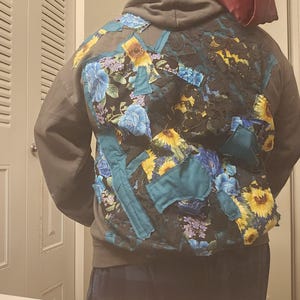 Peut inclure: Un sweat à capuche gris avec un motif patchwork au dos. Le patchwork présente des motifs floraux avec des tournesols, des fleurs bleues et des accents turquoise. Le design est un mélange de tissus et de textures.