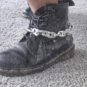Puede incluir: Un par de botas de cuero negro con costuras amarillas y un brazalete de tobillo de cadena plateada.