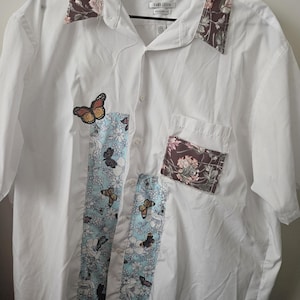 Op de afbeelding: Een wit overhemd met korte mouwen, knoopsluiting en een bruine kraag en borstzak met bloemenprint. Een verticaal paneel van lichtblauwe stof met een witte bloemen- en vlinderprint loopt over de voorkant. Het label bevat de tekst Van Heusen.