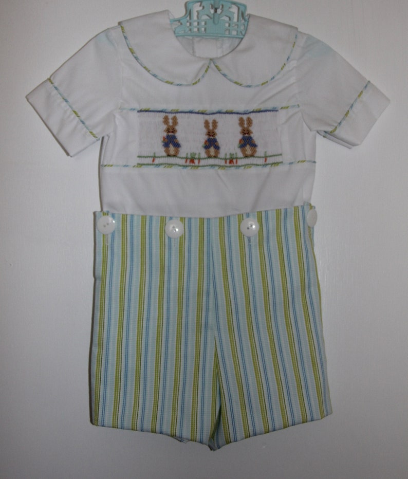 May include: A white baby outfit with a Peter Pan collar and short sleeves. The top features a cross-stitched design of three bunnies. The shorts are striped in blue and green, with large white buttons on the sides. A light blue hanger holds the outfit.