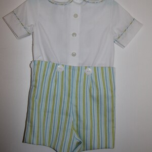 May include: A white short-sleeved romper set with a Peter Pan collar. The top has three buttons down the front. The shorts are striped in blue and green. The collar and sleeves have a thin trim of blue and green.