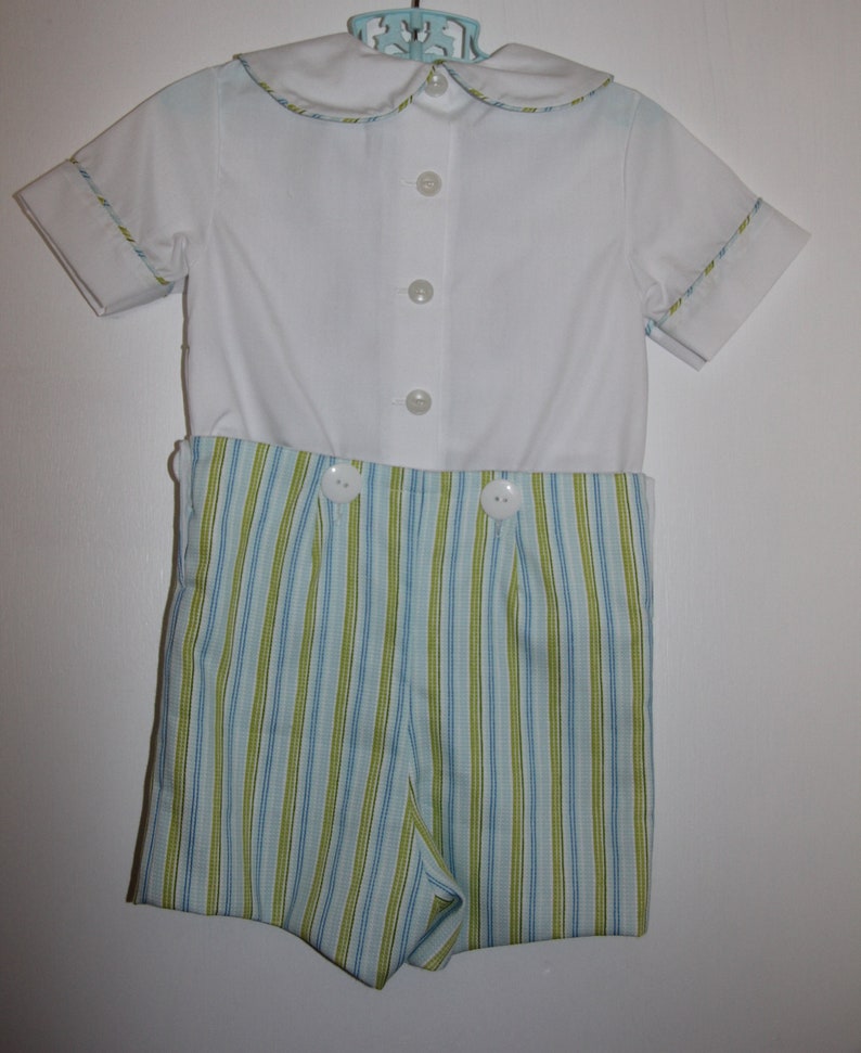 May include: A white short-sleeved romper set with a Peter Pan collar. The top has three buttons down the front. The shorts are striped in blue and green. The collar and sleeves have a thin trim of blue and green.
