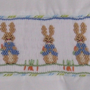 May include: White fabric with a smocked panel featuring three embroidered bunnies wearing blue jackets and holding carrots. The panel is bordered with blue and green trim, suggesting a children's item.