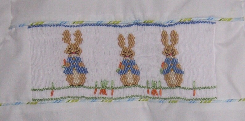 May include: White fabric with a smocked panel featuring three embroidered bunnies wearing blue jackets and holding carrots. The panel is bordered with blue and green trim, suggesting a children's item.