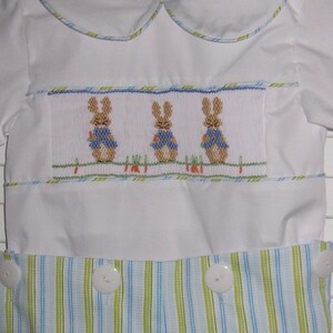 May include: White romper with a Peter Rabbit design. The romper features a smocked panel with three embroidered bunnies wearing blue vests. The bottom half has blue and green stripes, and the outfit has white buttons.