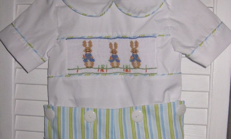 May include: White romper with a Peter Rabbit design. The romper features a smocked panel with three embroidered bunnies wearing blue vests. The bottom half has blue and green stripes, and the outfit has white buttons.