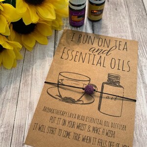 May include: A brown card with the text "I run on tea and essential oils" and a drawing of a teacup and a bottle of essential oil. A purple lava bead bracelet is wrapped around the card and the teacup. The text "Aromatherapy lava bead essential oil diffuser. Put it on your wrist & make a wish. It will start to come true when it falls off of you! Alyssa Ashley Designs" is printed on the card.