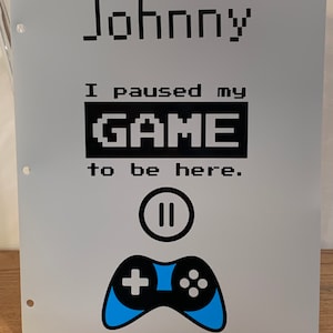 May include: A gray binder with the text "Johnny I paused my GAME to be here." in black. A blue and black video game controller is below the text with a pause button symbol above it.