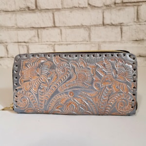 May include: A silver-grey leather wallet with a floral embossed design. The wallet has a zip closure and a gold-coloured zip pull. The floral pattern is in a lighter, orange-brown colour, creating a two-tone effect. The wallet is rectangular.