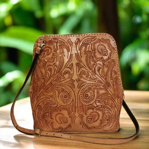 May include: A tan leather shoulder bag with intricate floral tooling. The bag has a long adjustable strap and a flap closure.