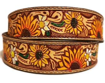 May include: Two stacked, hand-tooled leather belts with a sunflower design. The belts feature yellow, orange, and white sunflowers with green leaves and brown accents. The background is a light tan color.