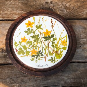 Könnte beinhalten: Ein rundes Käseboard aus Holz mit einem weißen Keramikeinsatz, der mit einem floralen Muster aus gelben Blumen und grünen Blättern verziert ist. Der Text "Goodwood®" ist auf den Keramikeinsatz gedruckt.