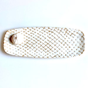 May include: White ceramic serving tray with a gold geometric pattern. The tray is oval shaped and has a small indentation on one end.