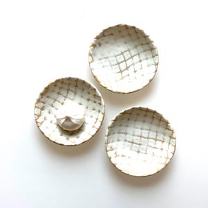 May include: Three small white ceramic dishes with a gold grid pattern. One dish has a single clove of garlic in the center.